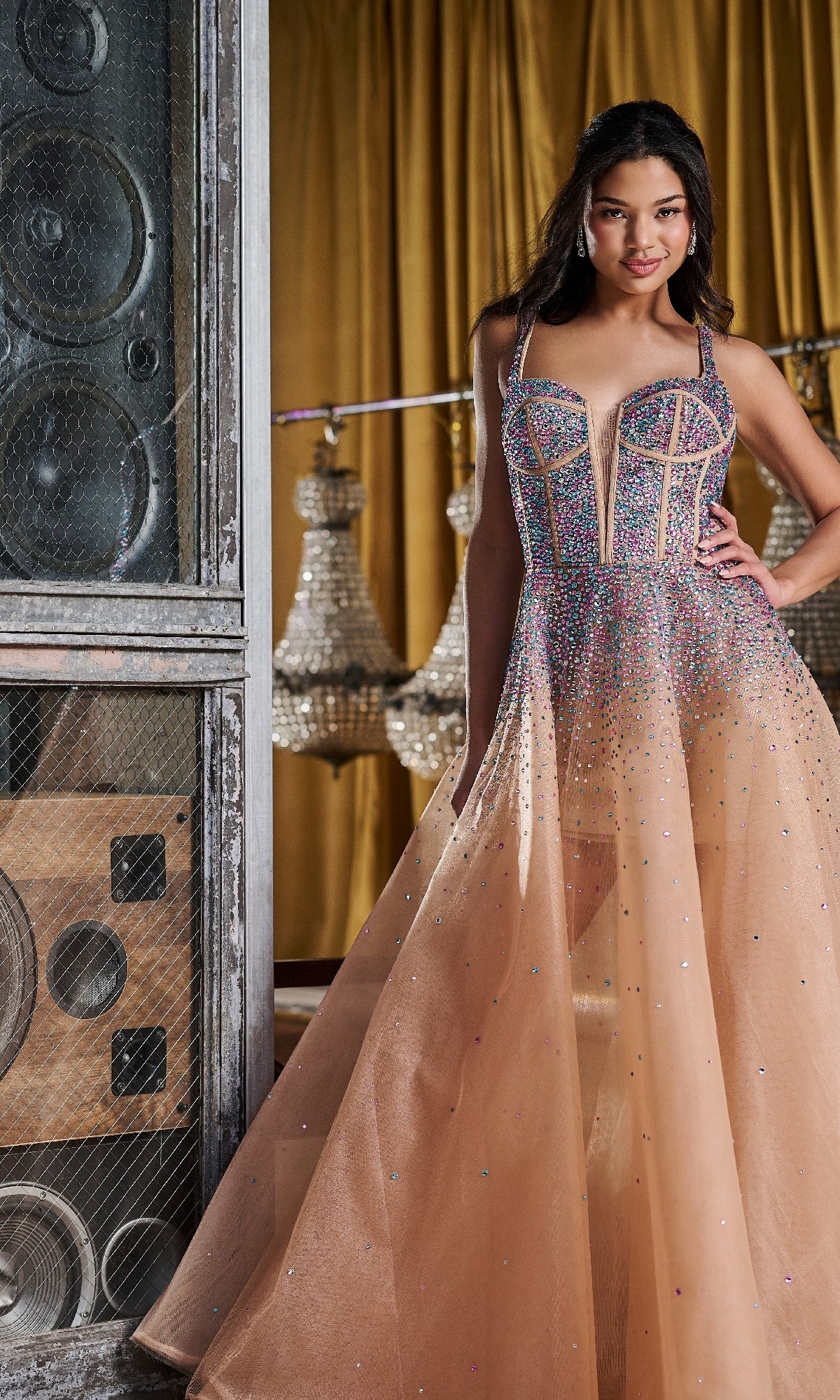 A woman poses indoors near large speakers and chandeliers, wearing the Tiffany 16206 long prom dress—a sparkling peach gown with crystal embellishments—her hand resting on her hip.